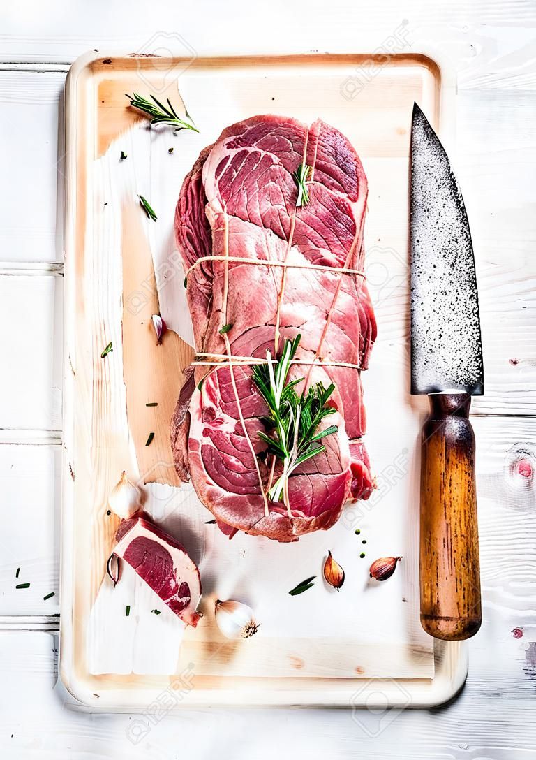 Raw uncooked roastbeef meat cut with rosemary, thyme and garlic and butcher knife on old white painted wooden background, top view, vertical composition