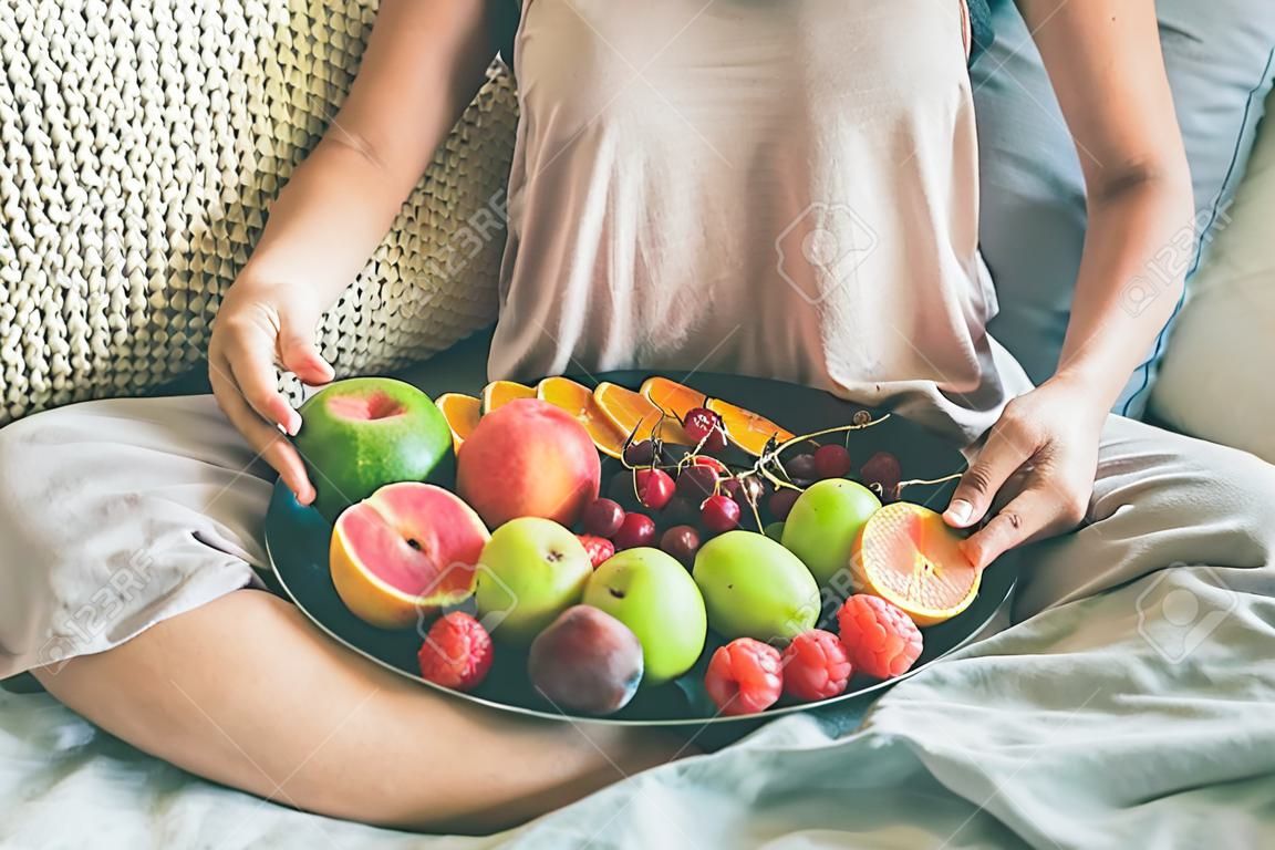Desayuno sano sano crudo de la consumición del vegano del verano en concepto de la cama. Chica joven que lleva ropa casera coloreada pastel que sienta y que sostiene la bandeja llena de fruta estacional fresca