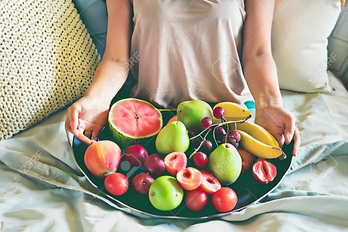 Summer healthy raw vegan clean eating breakfast in bed concept. Young girl wearing pastel colored home clothes sitting and holding tray full of fresh seasonal fruit