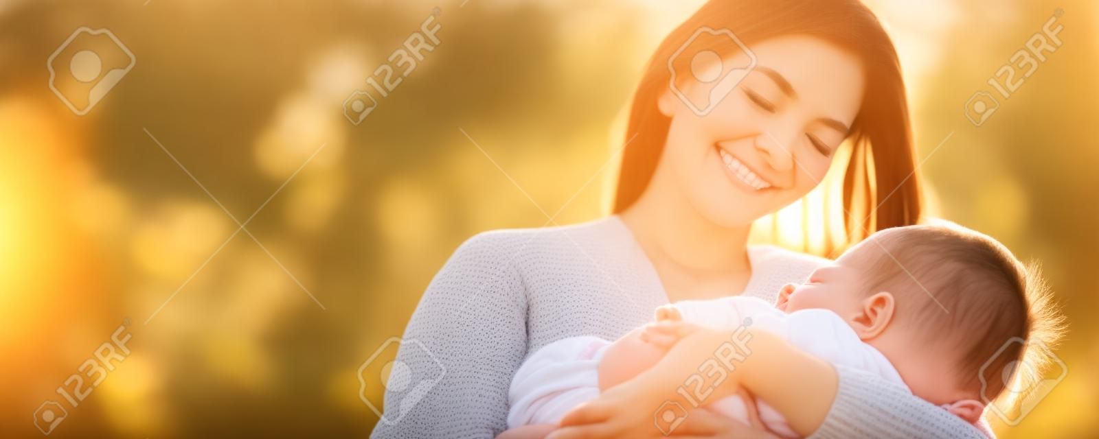 Ein herzerwärmender Muttertagsmoment, festgehalten im Park bei Sonnenuntergang. Eine schöne Mutter wiegt ihr schlafendes Baby, sonnt sich in der friedlichen Natur und lächelt ihr Neugeborenes liebevoll an. Familienbetreuung vom Feinsten
