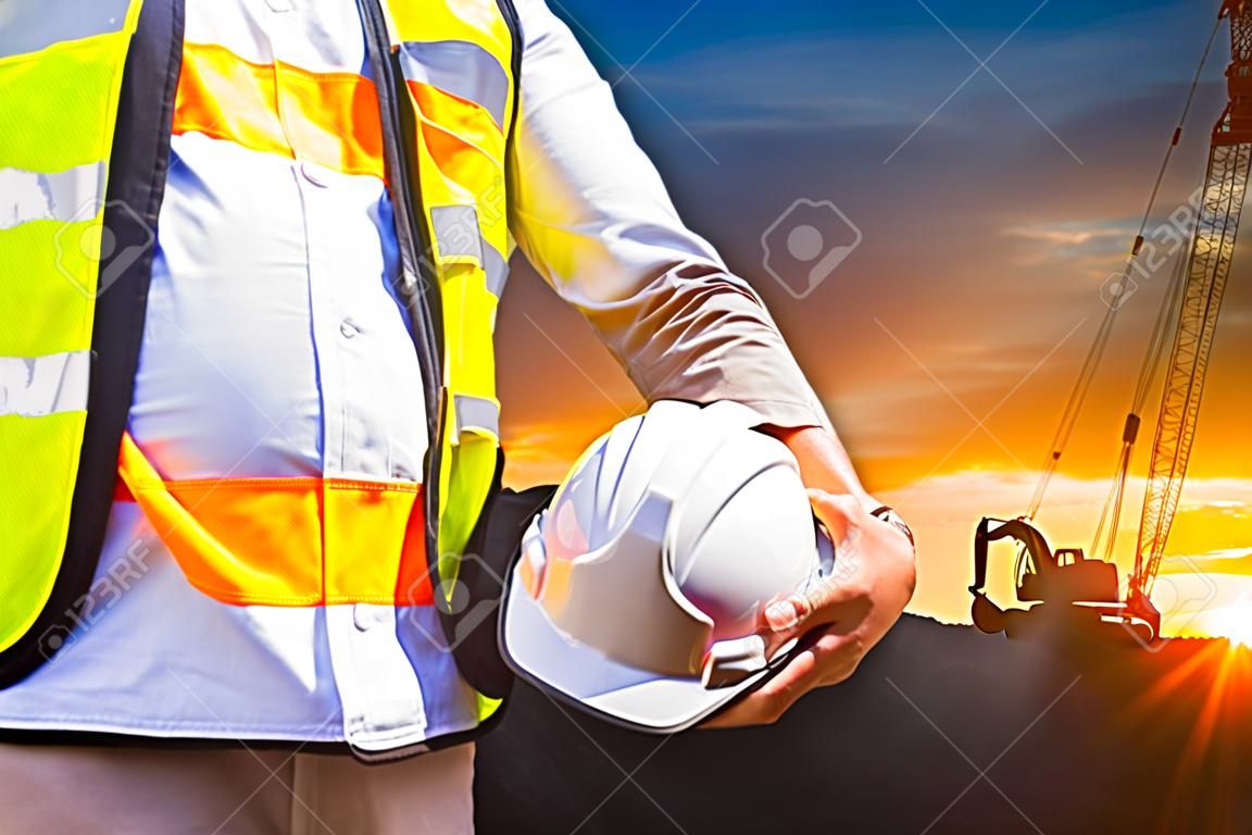 Ingenieur oder Sicherheitsbeauftragter harten Hut mit Bagger Maschine auf der Baustelle auf Sonnenuntergang Zeit hält, ist Hintergrund.