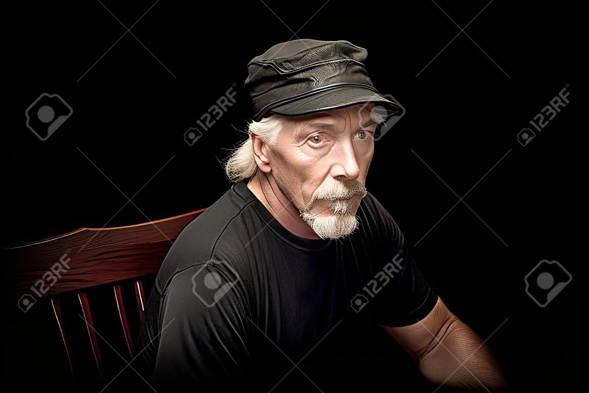 An older white biker male  with beardsitting in a dark room wearing black t shirt and leather cap, looking serious at viewer.