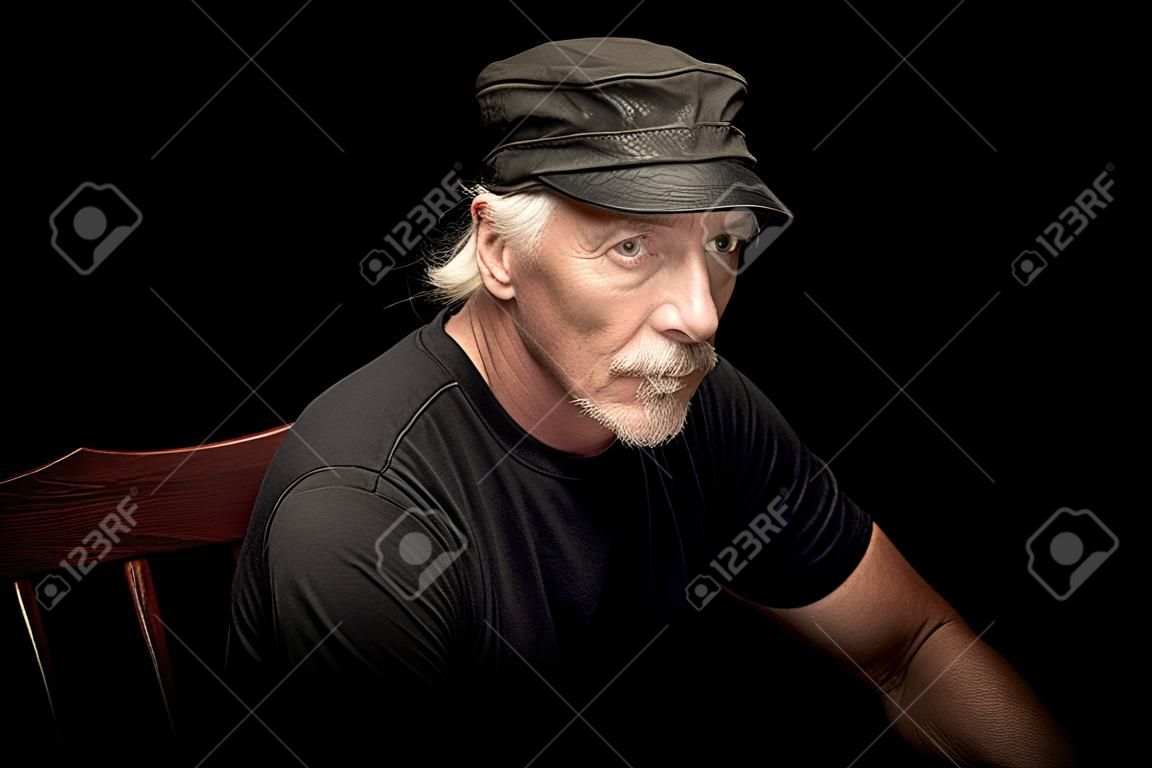 An older white biker male  with beardsitting in a dark room wearing black t shirt and leather cap, looking serious at viewer.