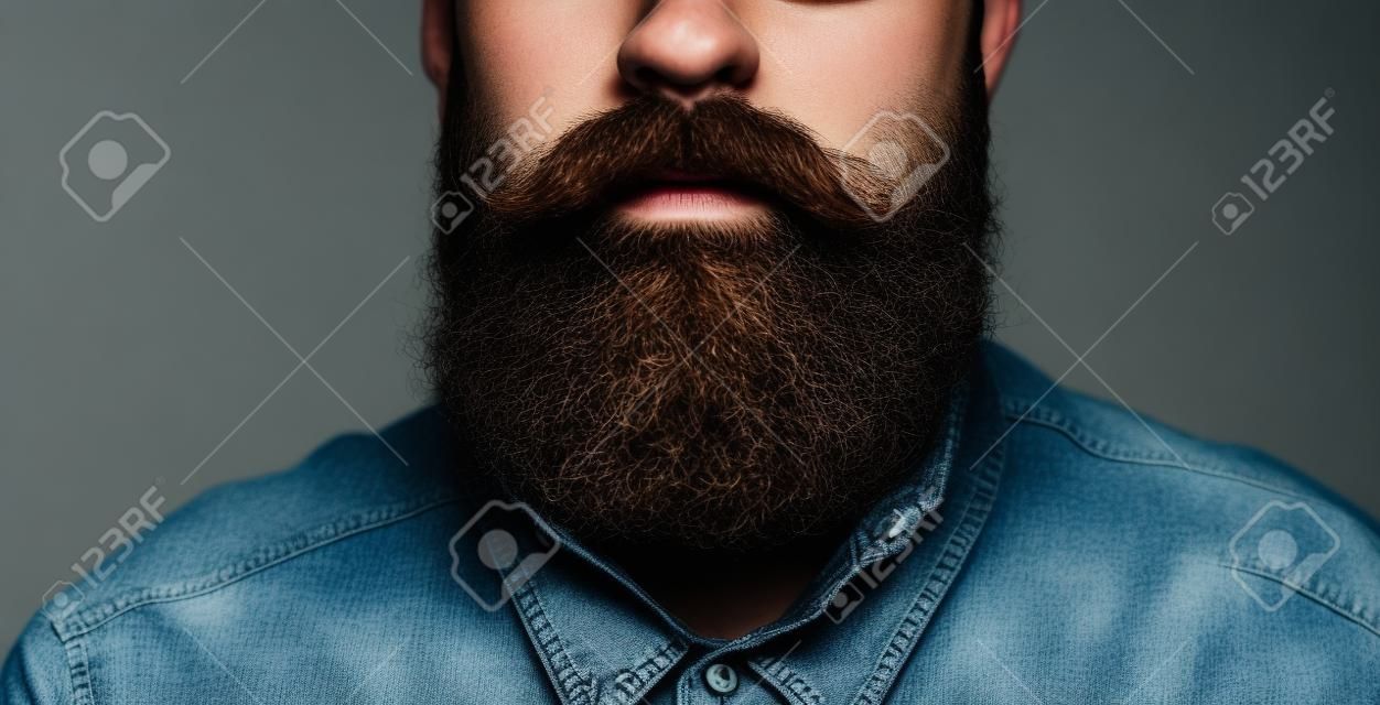 Atrybuty wyglądu mężczyzny hipster: wąsy i broda z bliska. Portret brodatego mężczyzny w niebieskiej dżinsowej koszuli na czarnym tle