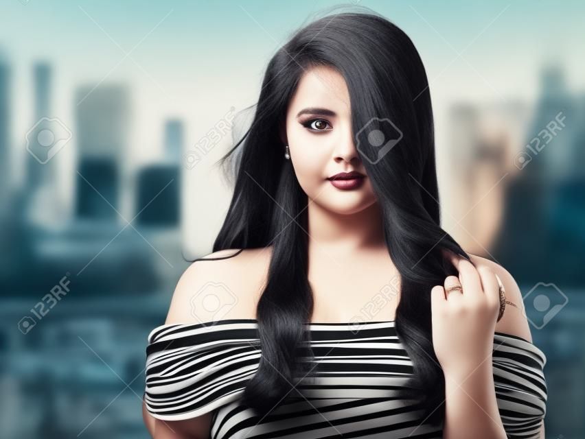 Portrait of young beautiful curvy woman plus size in striped top and black skirt over blurred city background