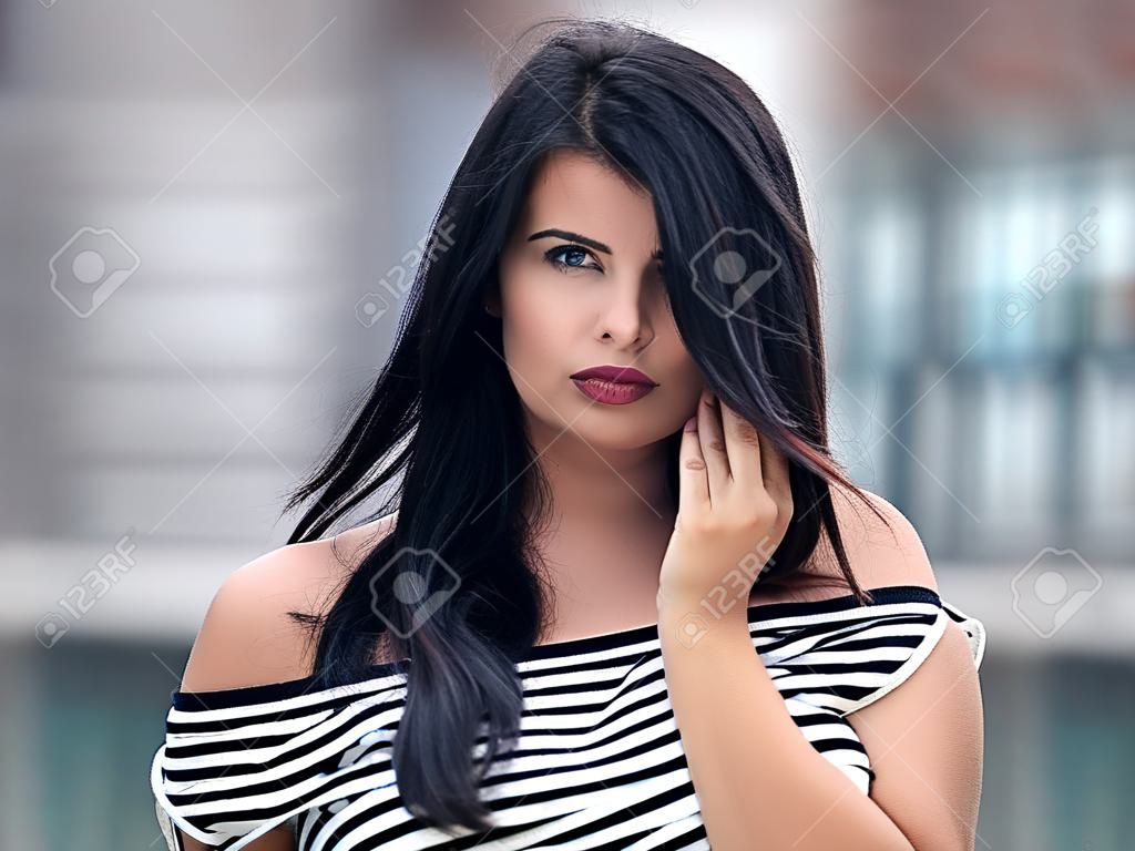 Portrait of young beautiful curvy woman plus size in striped top and black skirt over blurred city background