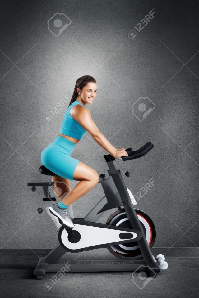 운동복을 입은 아름다운 운동복 젊은 브루네트 여성이 회색 벽을 배경으로 체육관에서 사이클을 타고 있습니다.