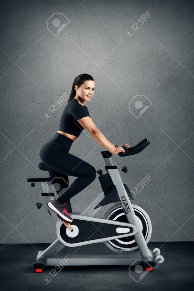 Una hermosa joven morena atlética con ropa deportiva entrena en un ciclo en el gimnasio con el telón de fondo de una pared gris.