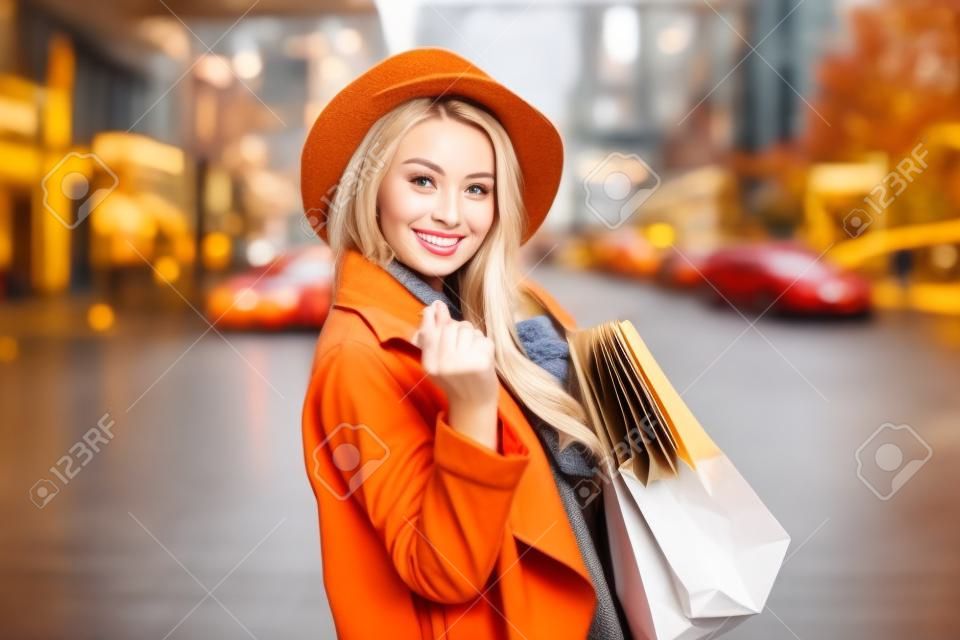 Cute attractive shopaholic woman in autumn coat and hat holding many paper shopping bags in the city