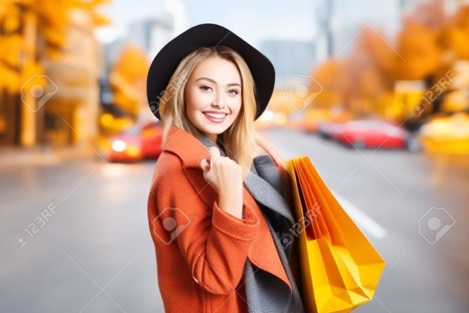 도시에서 많은 종이 쇼핑백을 들고 있는 가을 코트와 모자를 쓴 귀엽고 매력적인 쇼핑 중독 여성