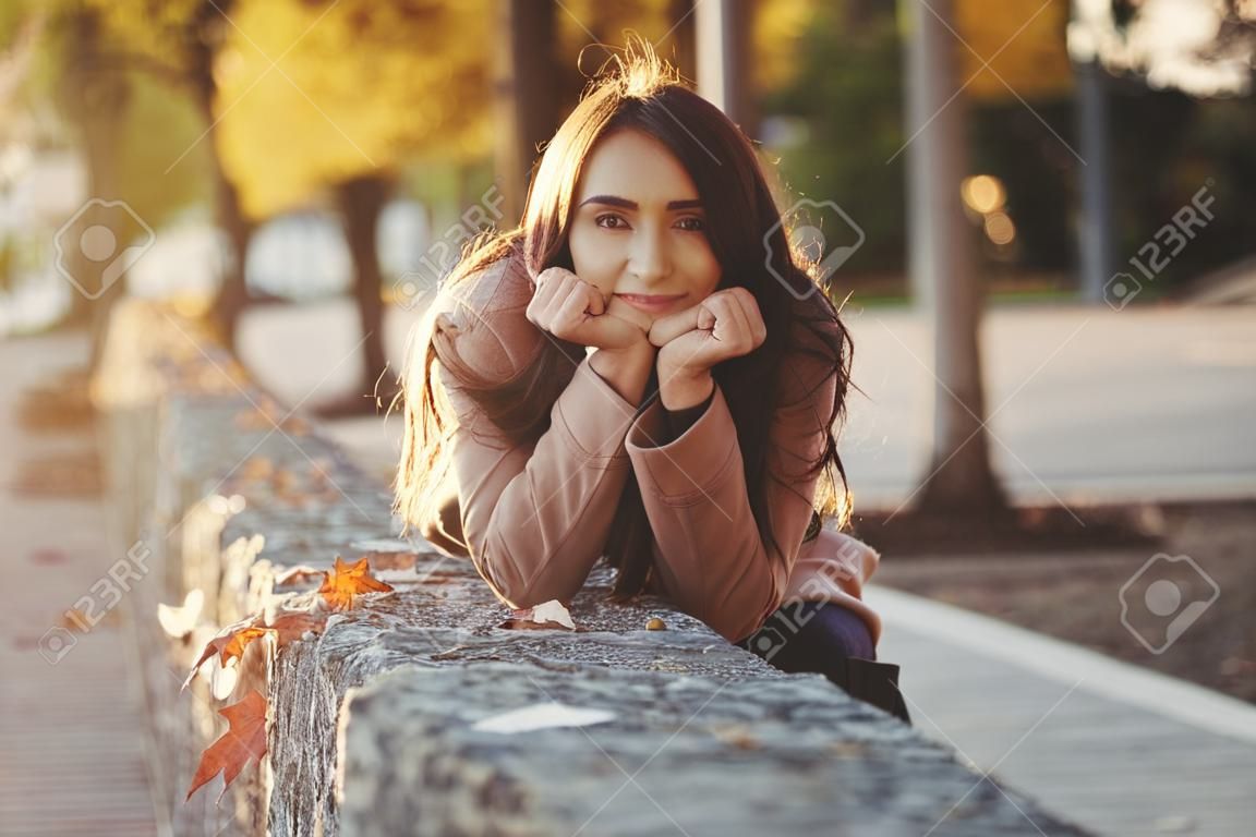 Jeune et jolie fille brune s'appuya sur le mur avec ses deux mains sous le menton, portant un manteau décontracté avec un parc ensoleillé d'automne sur le fond flou avec un espace de copie.