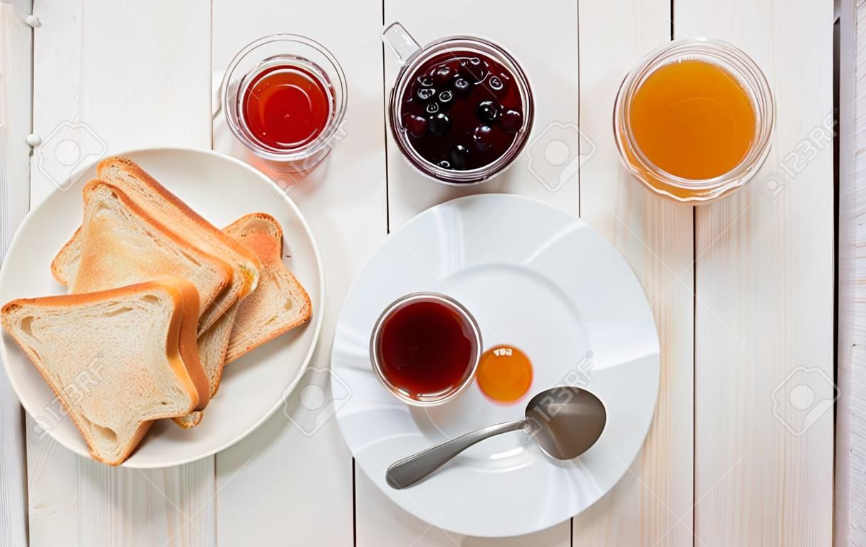Eine weiße Schale schwarzer Tee mit Sandwichen oder Toast mit Erdbeer-, Korinthen- und Aprikosengelee oder Stau auf weißem Holztisch, Draufsicht, flache Lage, Frühstückskonzept mit Kopienraum
