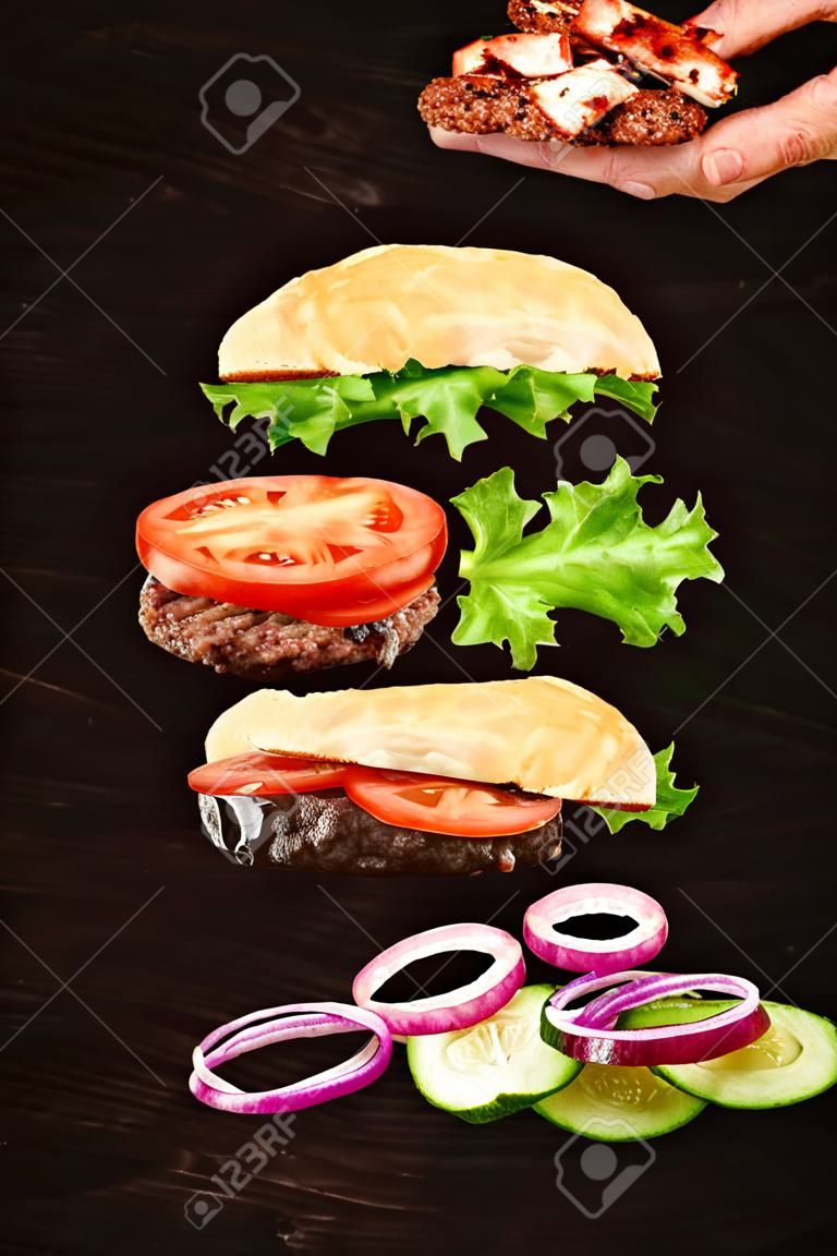 Floating burger isolated on black wooden background in men hands. Ingredients of a delicious burger with ground beef patty, lettuce, bacon, onions, tomatoes and cucumbers