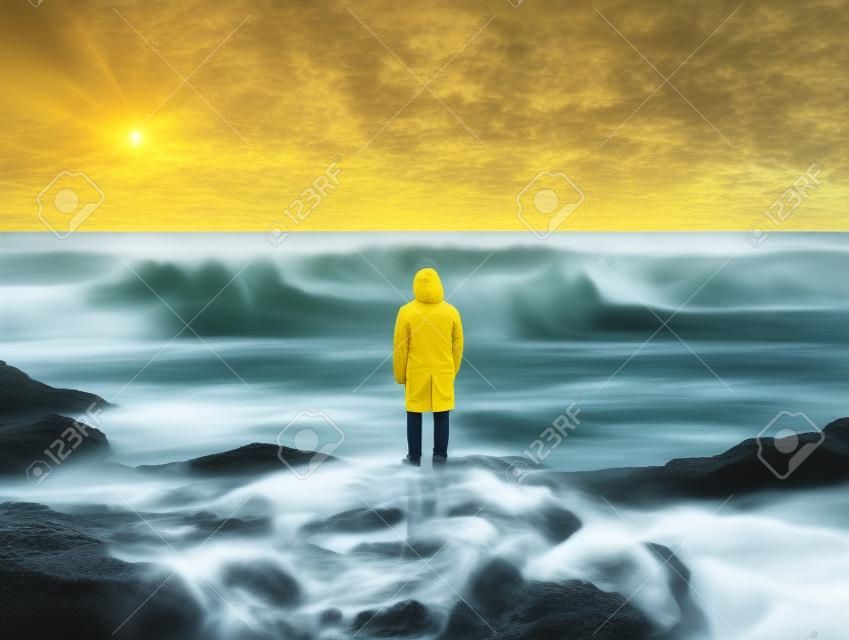 A person in a yellow raincoat standing on rocks in front of the ocean. AI generative image.