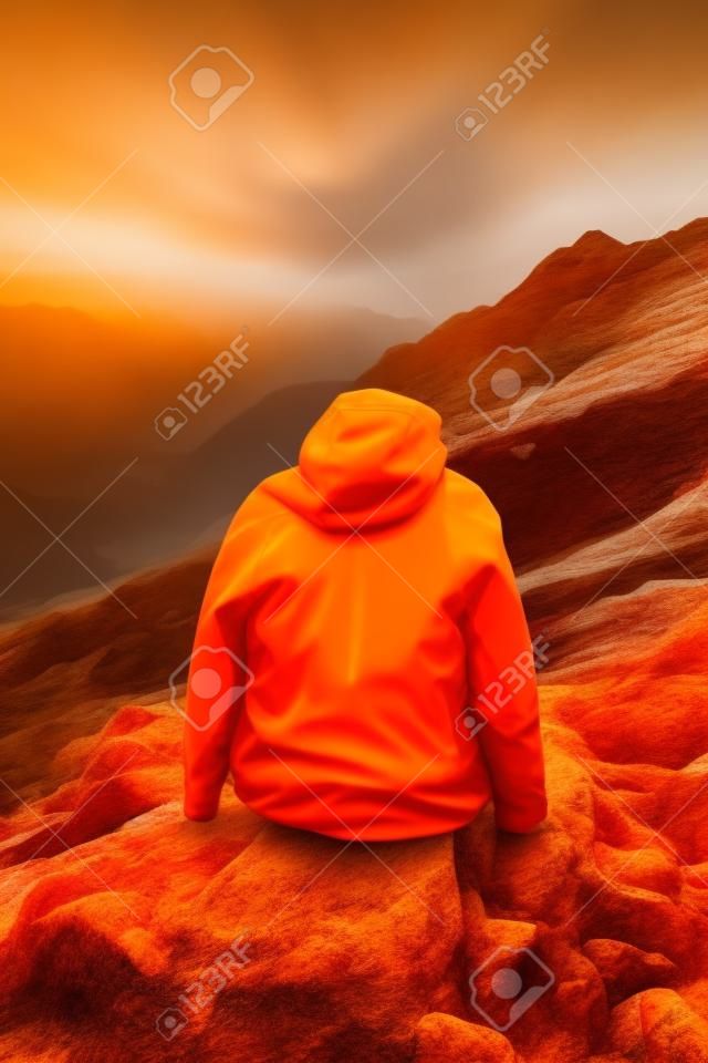 Eine Person in einer orangefarbenen Jacke sitzt auf einem Felsen. Generatives KI-Bild.