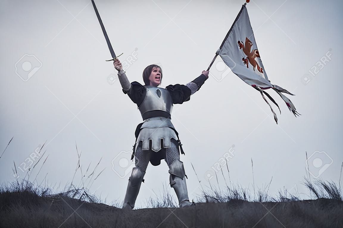 La ragazza nell'immagine del guerriero sta in armatura ed emette un grido di battaglia con la spada sollevata e la bandiera tra le mani su sfondo di cielo ed erba secca.