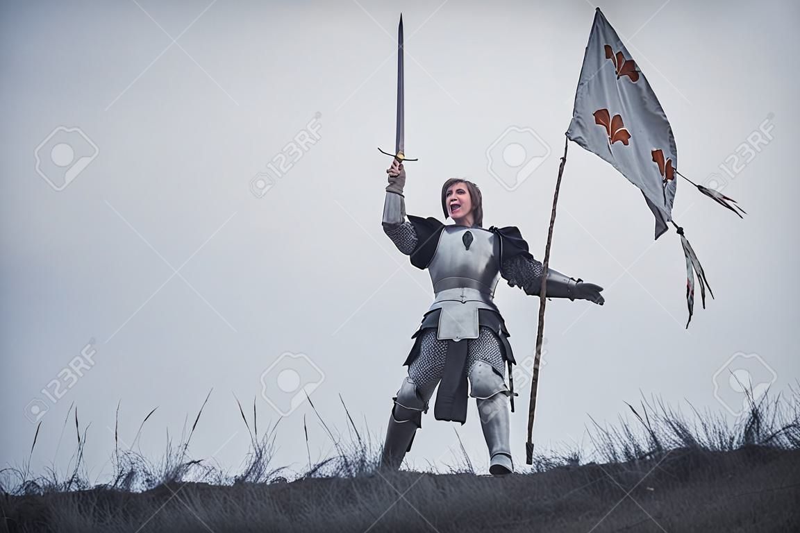Mädchen im Bild des Kriegers steht in der Rüstung und gibt Kampfschrei mit der angehobenen Klinge und Flagge in ihren Händen gegen Hintergrund des Himmels und des trockenen Grases aus.