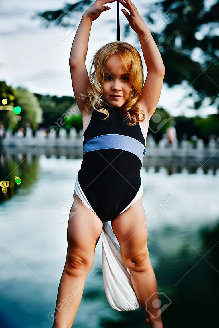 Child girl aerialist performs acrobatic tricks on hanging aerial silk against background of river, sky and trees.
