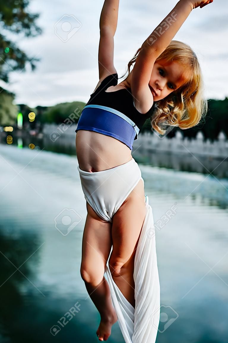 Child girl aerialist performs acrobatic tricks on hanging aerial silk against background of river, sky and trees.