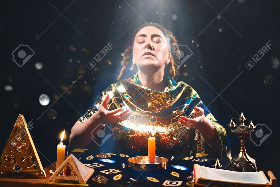 Astrology and divination. Portrait of a fortune teller who reads by candle, eyes closed in ecstasy. Black background. There are magic items on the table.