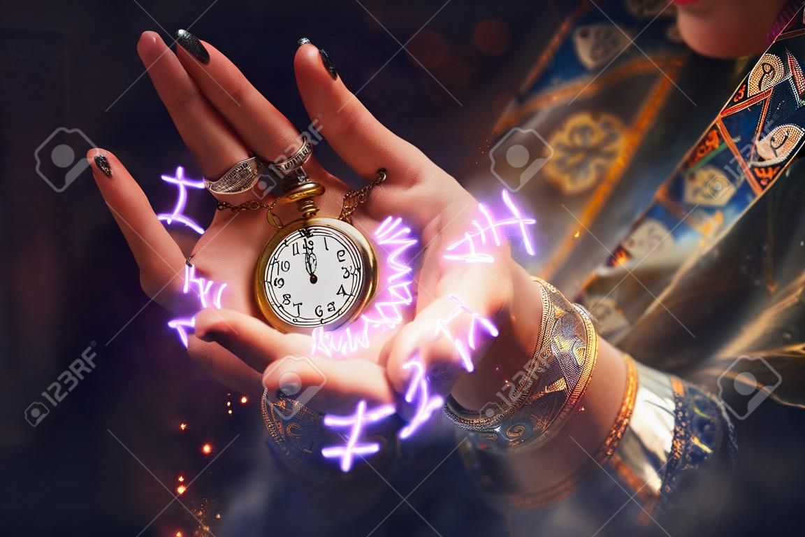 The fortune teller holds a watch on a chain in her hands and conjures over it. A luminous zodiac circle is depicted around the clock. The concept of divination, astrology and predicting the future.