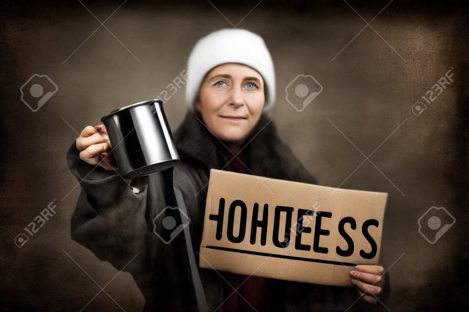 Una mujer caucásica sucia y desenfocada sostiene un cartel de cartón con el texto sin hogar y una taza de acero. fondo oscuro El concepto de ayudar a las personas sin hogar y vagabundos.