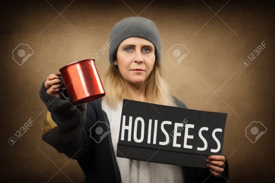 Una mujer caucásica sucia y desenfocada sostiene un cartel de cartón con el texto sin hogar y una taza de acero. fondo oscuro El concepto de ayudar a las personas sin hogar y vagabundos.