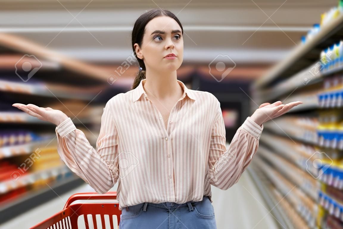 Retrato de surpresa jovem mulher caucasiana segura carrinho de compras e espalha os braços para os lados. prateleiras de loja de mercearia e corredor em segundo plano. Conceito de escolha e compras.