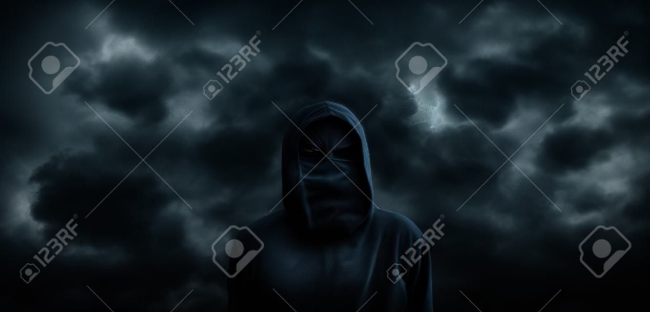 Spooky hooded person with obscured face against the dark dramatic sky