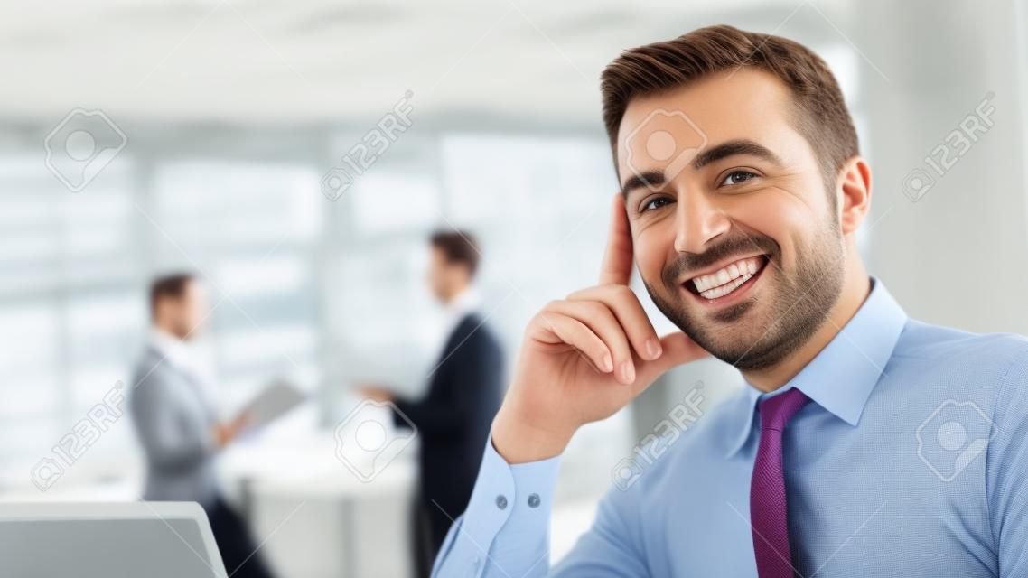Confident handsome businessman sitting at office desk and smiling at camera, office interior and business people standing on background, selective focus
