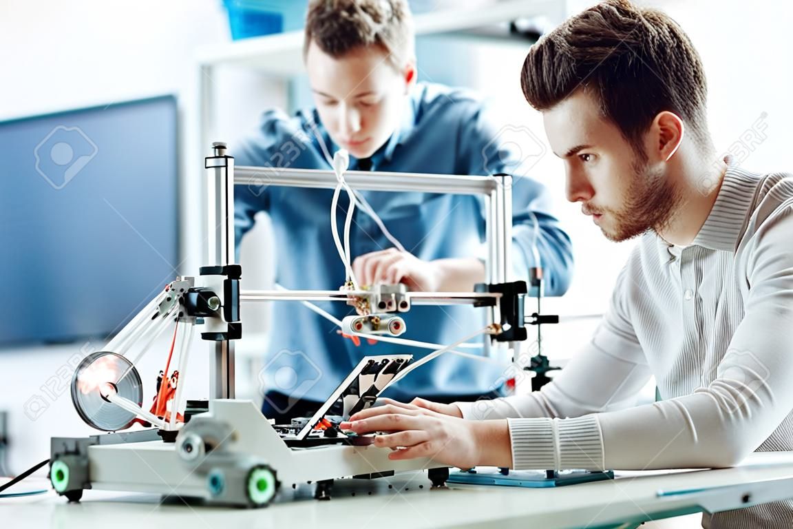 Estudantes de engenharia que trabalham no laboratório, um aluno está ajustando os componentes de uma impressora 3D, o outro em primeiro plano está usando um laptop