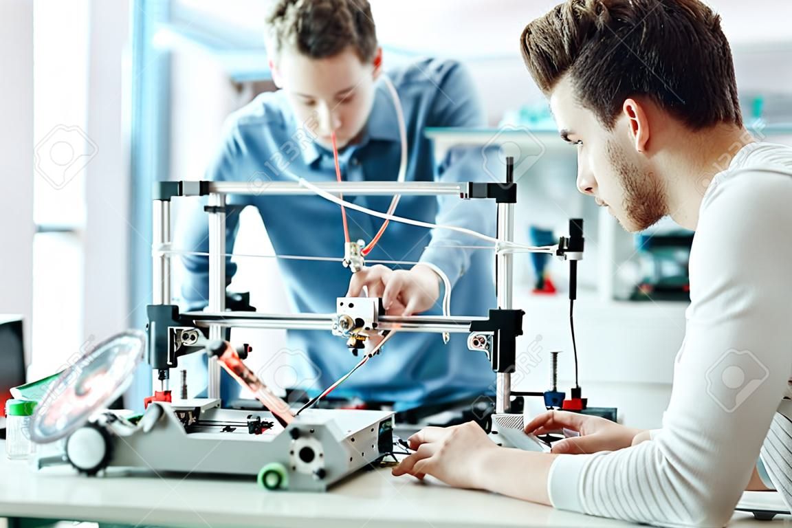 Bir öğrenci 3D Yazıcının parçalarını ayarlama olduğu laboratuvarda çalışan Mühendisliği öğrencileri, ön planda diğeri laptop kullanıyor