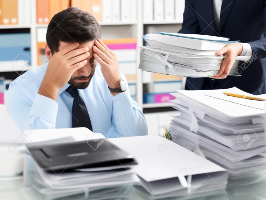 Stressed businessman working at office desk and overloaded with work, the desktop is covered with paperwork, his secretary is bringing more files