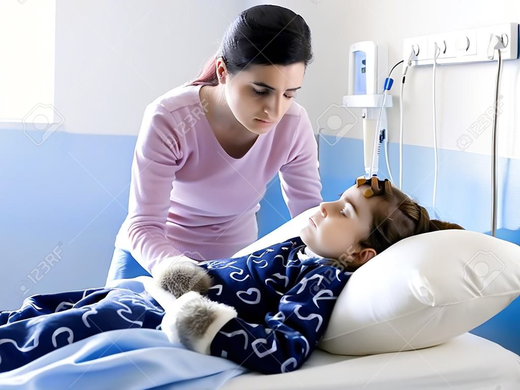 Young woman comforting her sick child lying in bed at the hospital, she is touching her head, pediatrics and healthcare concept