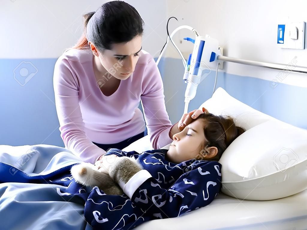 Young woman comforting her sick child lying in bed at the hospital, she is touching her head, pediatrics and healthcare concept