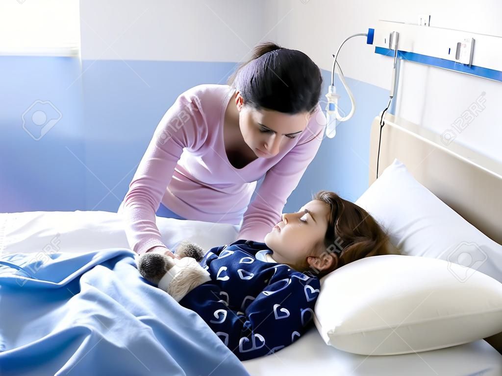 Giovane donna che conforta il suo bambino malato sdraiato a letto in ospedale, si sta toccando la testa, la pediatria e il concetto di assistenza sanitaria