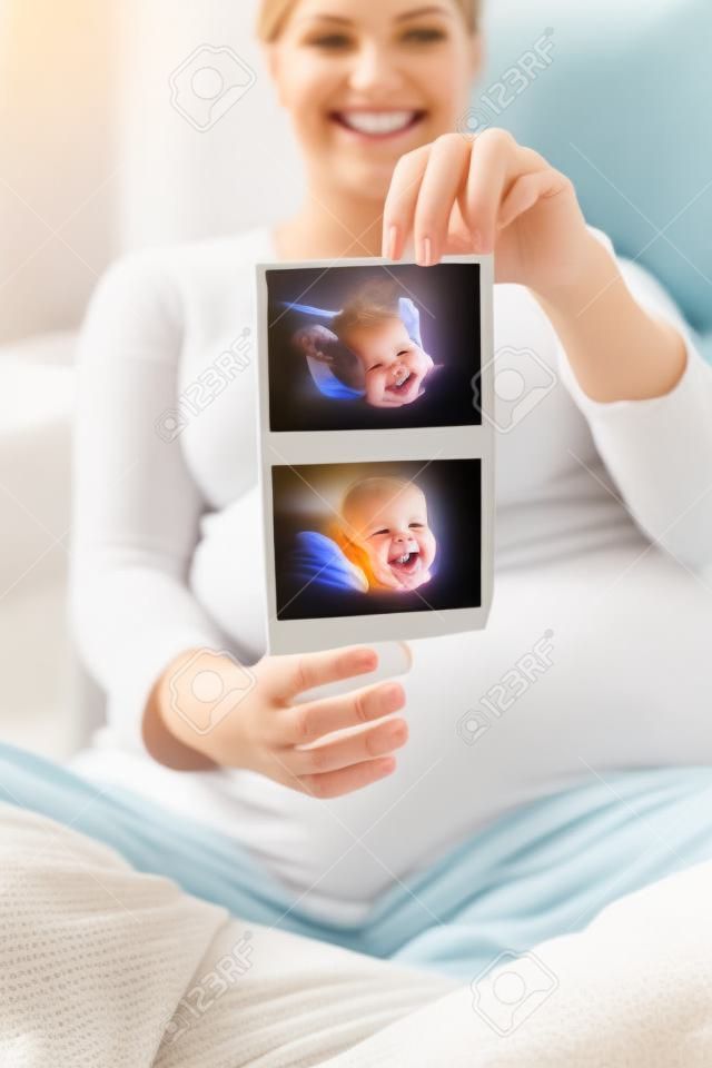 Happy smiling pregnant woman sitting on the sofa at home and showing ultrasound images of her baby