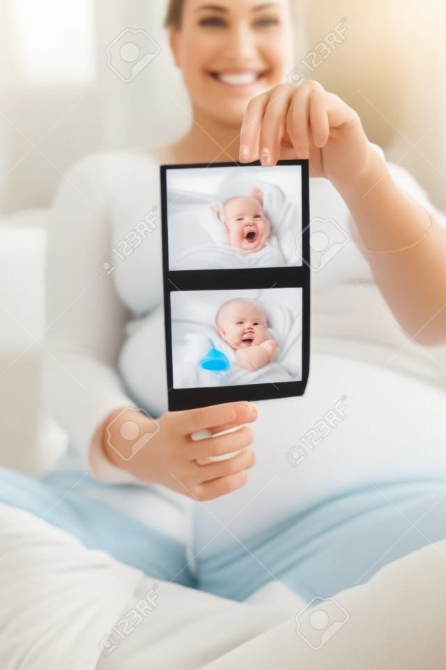 Happy smiling pregnant woman sitting on the sofa at home and showing ultrasound images of her baby