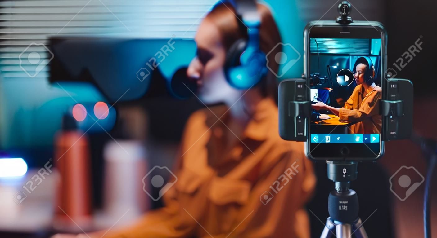 Mujer joven transmitiendo un video en vivo con su teléfono inteligente y hablando por un micrófono en el estudio
