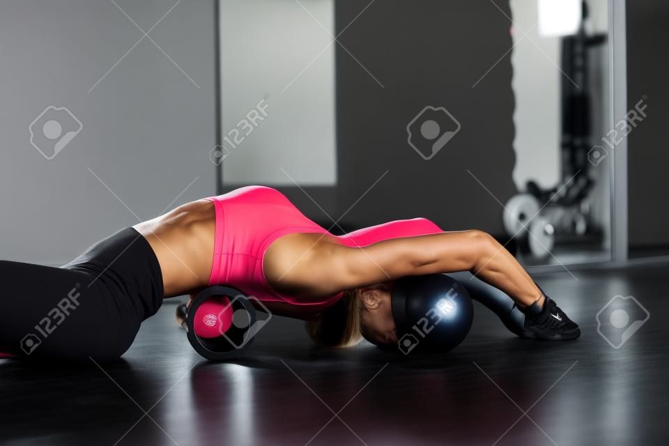 Fitnesstrainermädchen in schwarzen Leggings und T-Shirt im Trainingsraum beugt sich mit einer Massagerolle auf dem Boden im Loftraum