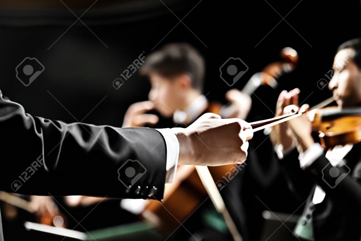 Dirigent leidt symfonieorkest met performers op achtergrond, handen close-up.