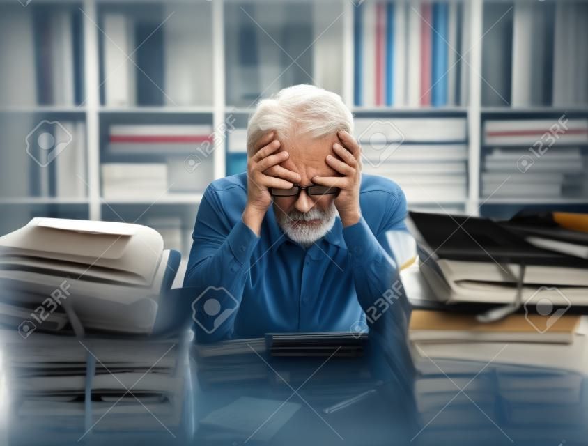Overloaded stressed senior office worker with head in hands, his desktop is filled with paperwork and files