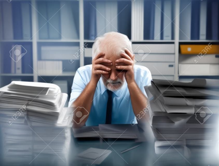 Overloaded stressed senior office worker with head in hands, his desktop is filled with paperwork and files
