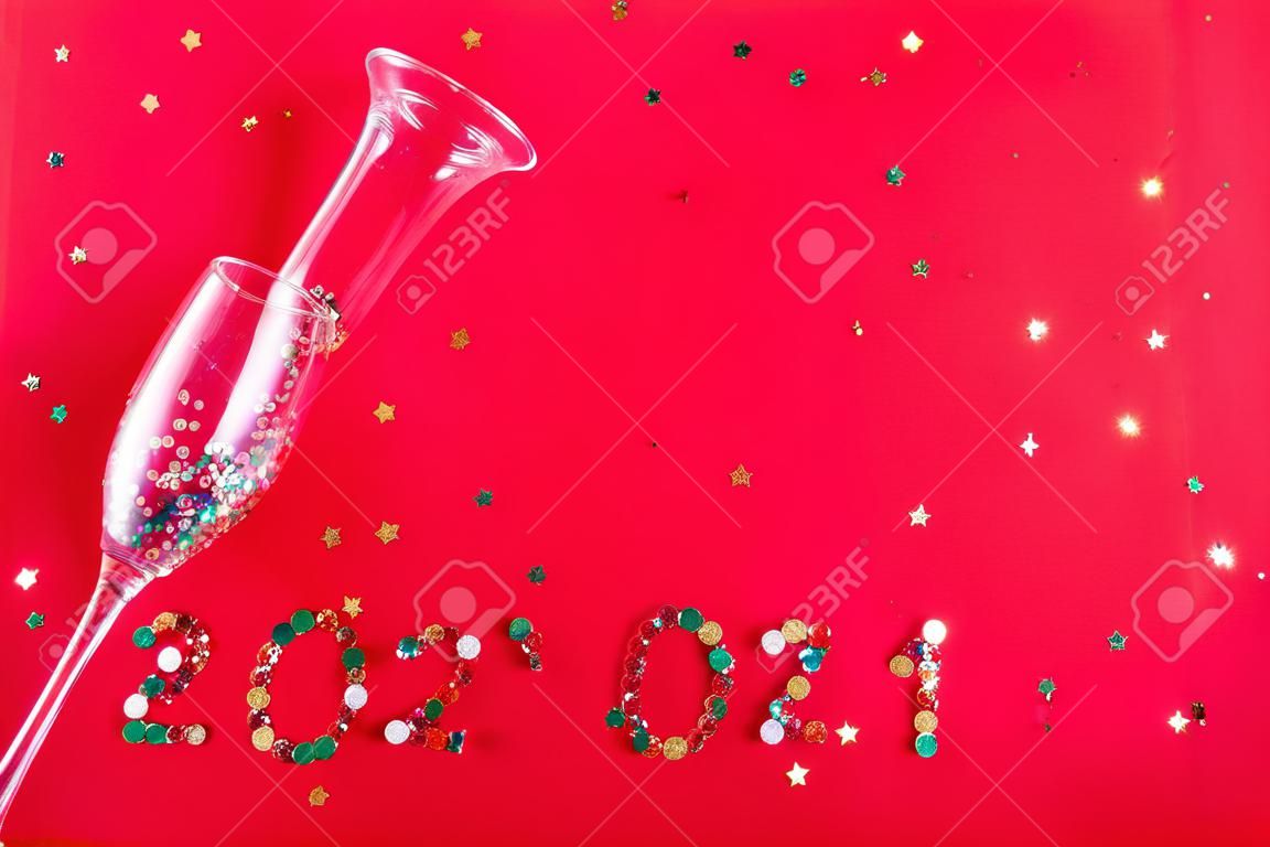 Bicchiere da champagne con paillettes a forma di stella sparse a forma di iscrizione 2021 su fondo rosso. Festa, Capodanno, concetto di celebrazione. Vista dall'alto. Disposizione piatta.