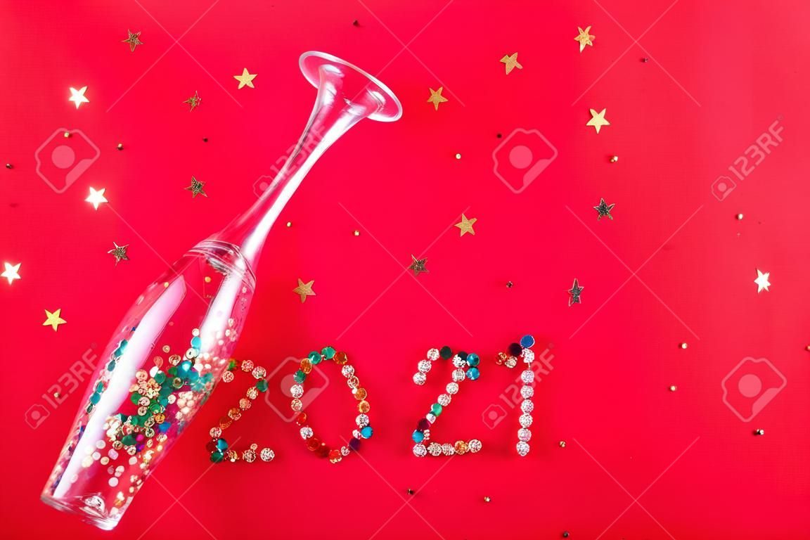 Champagnerglas mit sternförmigen Pailletten, die in Form der Aufschrift 2021 auf rotem Grund verstreut sind. Party, neues Jahr, Feierkonzept. Ansicht von oben. Flach liegen.