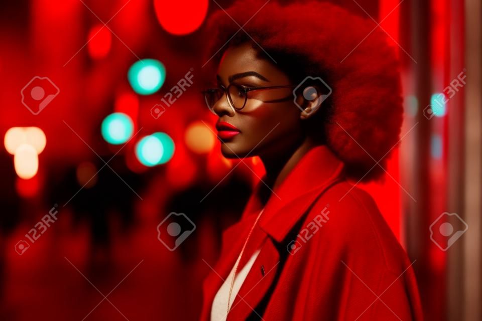 Fashionable african american woman in red coat and eyeglasses posing in the city at night.