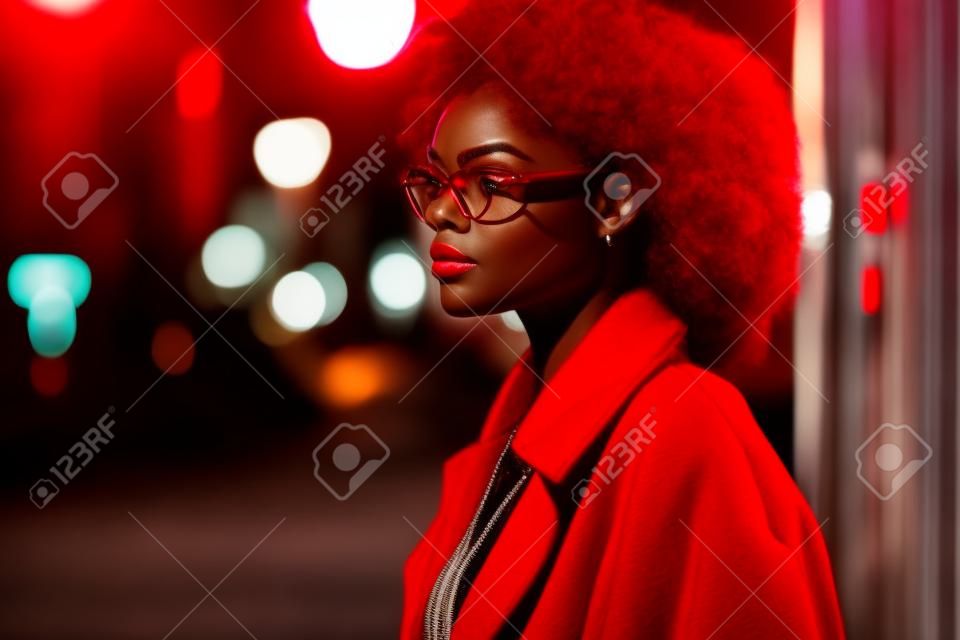 Fashionable african american woman in red coat and eyeglasses posing in the city at night.