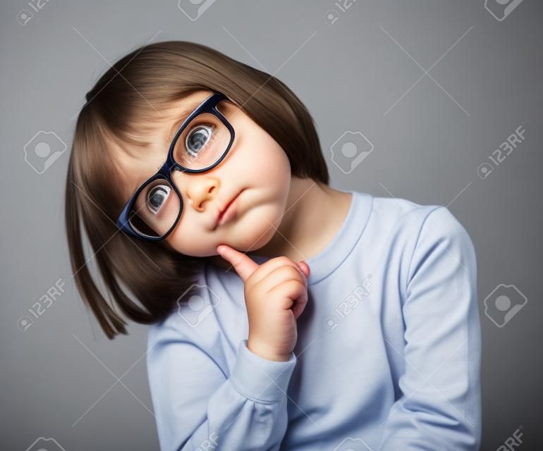 вдумчивый красивая маленькая девочка с серьезными очки держа ее голову, чтобы представить себе, думать, колеблясь или иметь интеллектуальную идею или решение, серый фон студийный