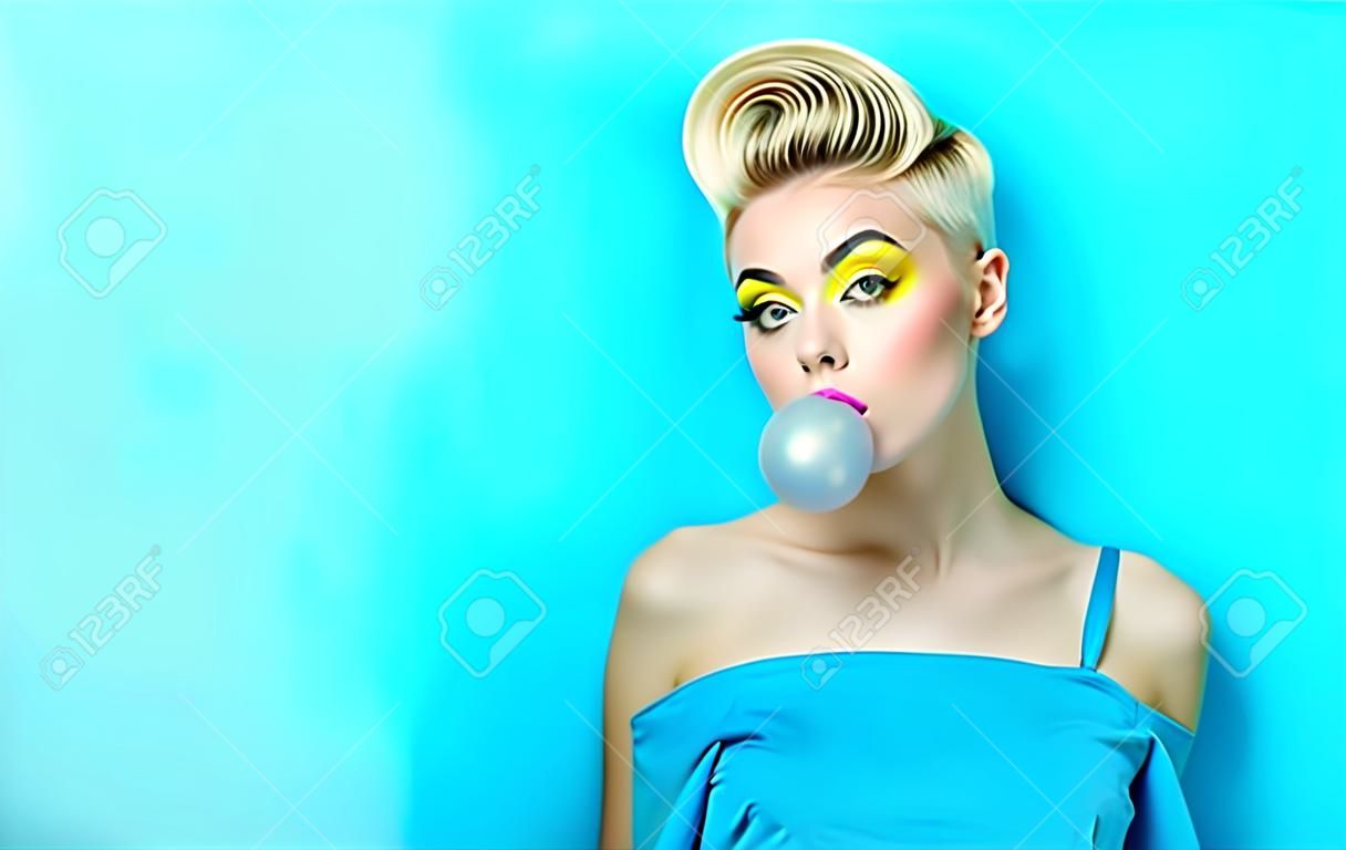 Модная девушка со стильной стрижкой надувает жевательную резинку. Девушка в студии на синем фоне. Лицо девушки с ярким макияжем и желтая с черными тенями на глазах.