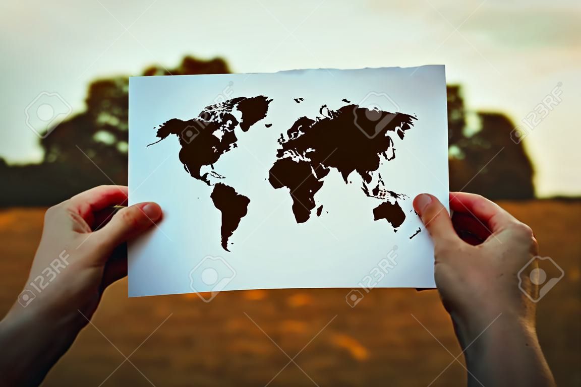 Climate change problem, destruction of nature concept. Human hands holding a paper sheet with world map icon over a dry grass field background. Global warming, solar radiation and pollution risk.